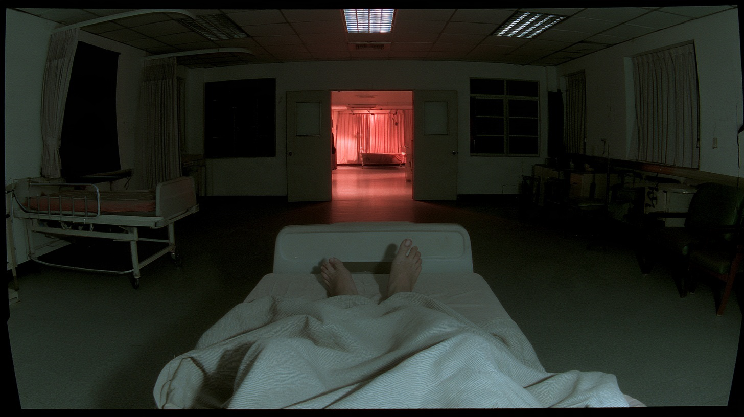 An empty green hospital ward seen from the bed, bare feet facing the open central doorway